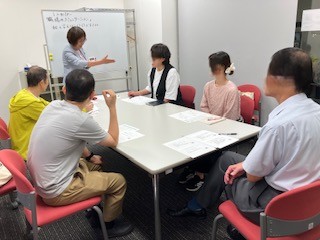 ホワイトボード前に立つ講師がテーブルを囲んだ5人のうちの1人に発言を促す